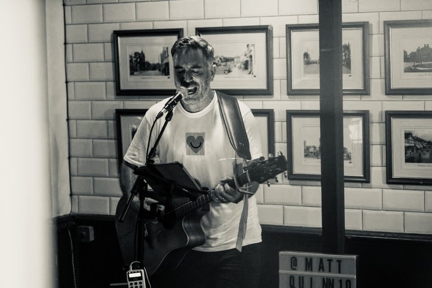 Black-and-white photograph of Matthew singing and playing acoustic guitar indoors