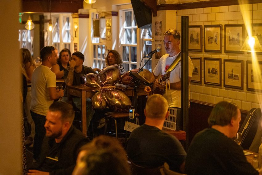 Matthew performing acoustic guitar in a warmly lit venue with guests nearby