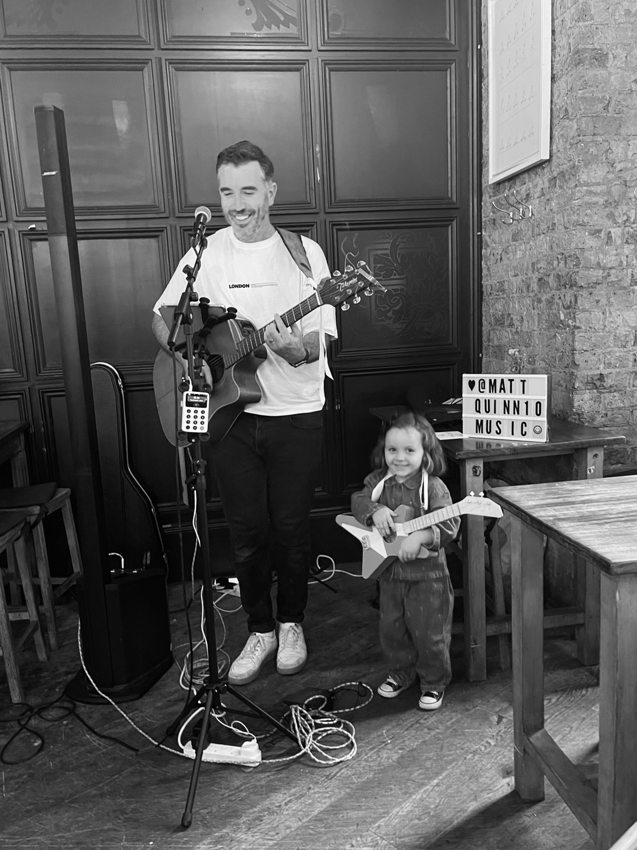 Black-and-white photograph of Matthew playing guitar beside a young guest holding a toy guitar