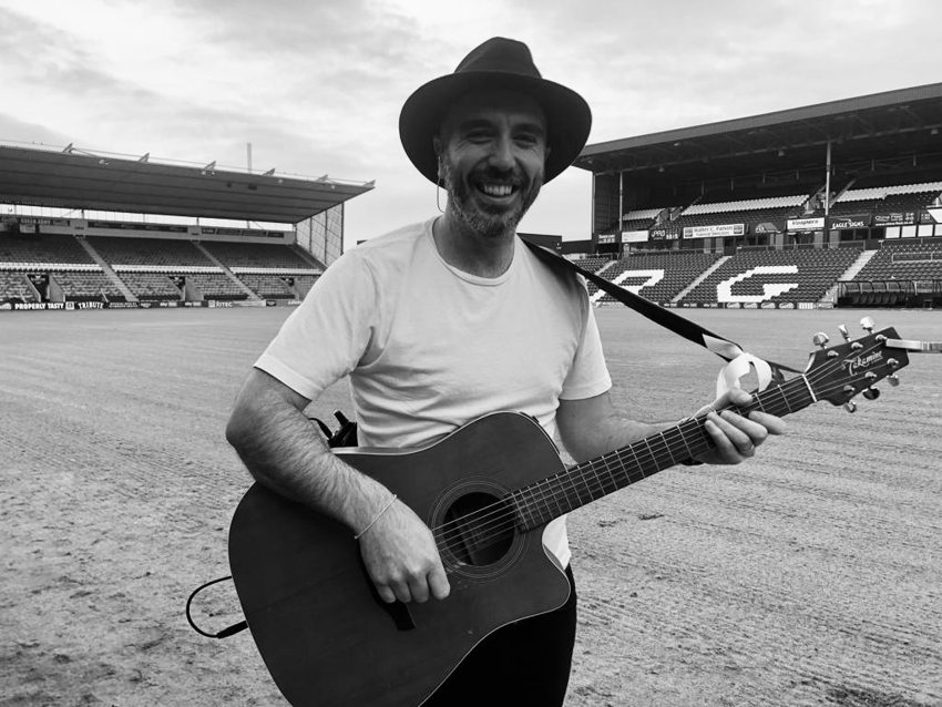 Black-and-white photograph of Matthew holding an acoustic guitar on a stadium pitch